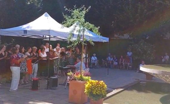 Der Chor VoiceNet singt und klatscht rhythmisch beim Singen im Hofgarten in Augsburg bei Sonnenschein und Regenbogen-Effekt umgeben von Natur und Publikum unter Pavillons